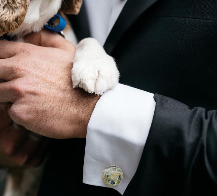Close-up of a person's hand holding a dog's paw, with a focus on the memorial cremation ash cufflink.